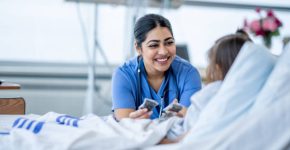 A female nurse of Middle Eastern decent sits at the edge of a hospital bed as she check in on her young patient. She is wearing blue scrubs and is attempting to cheer the young girl up.